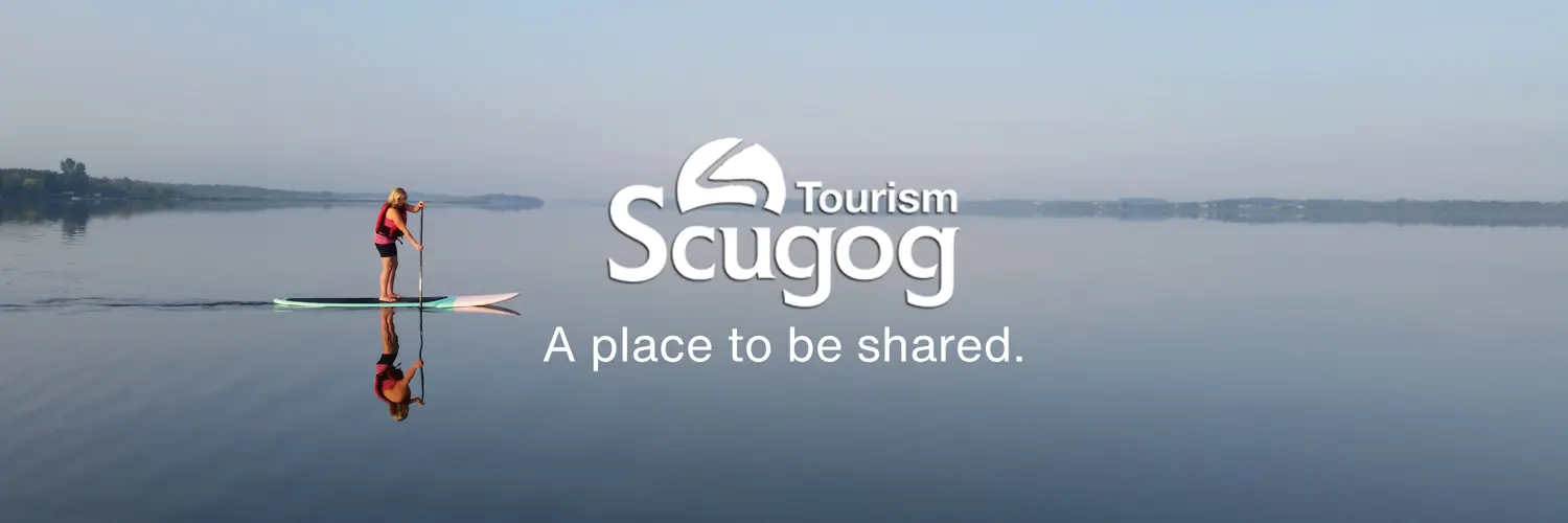 person on standup paddle board on Lake Scugog with overlaid Scugog Tourism logo and white text reading 'A place to be shared.'